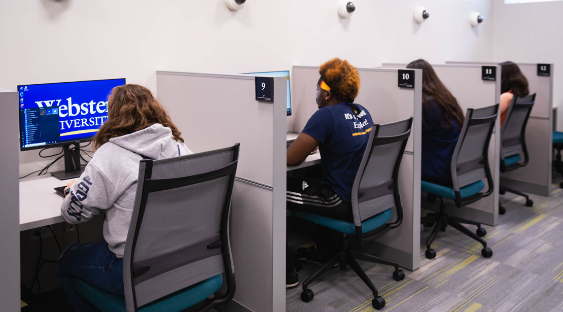 Four computer testing cubicles with computer monitors and dividers are in a row along the wall of the Reeg Academic Resource Center's testing center.  Each station has a camera on the wall above it, and a student occupies the wheeled chair in front of its monitor.