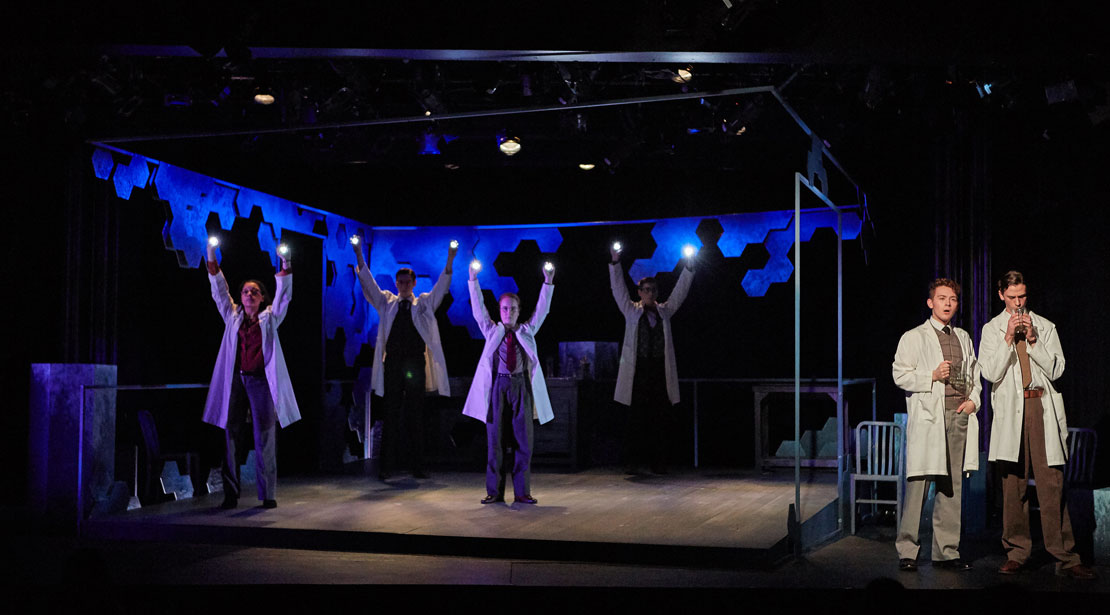 Whole set with four actors holding lights above their heads on the left and two actors wearing lab coats talking together on the right