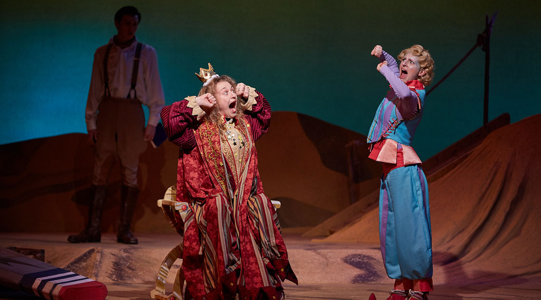An actor wearing a crown in red and gold robes with hands in fists next to head stands to the left of the actor in blue and purple whose arms are held the same way