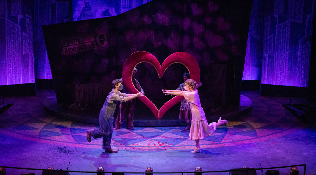 Two actors are facing each other, both standing on a single leg with the other bent behind them with their arms reaching for the other; there is a large heart in the background behind them
