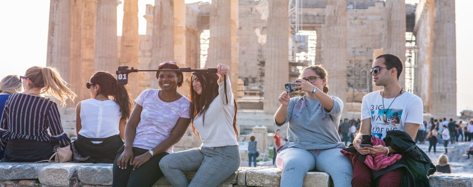 People sit on low stone wall taking selfies with Greek ruins behind them