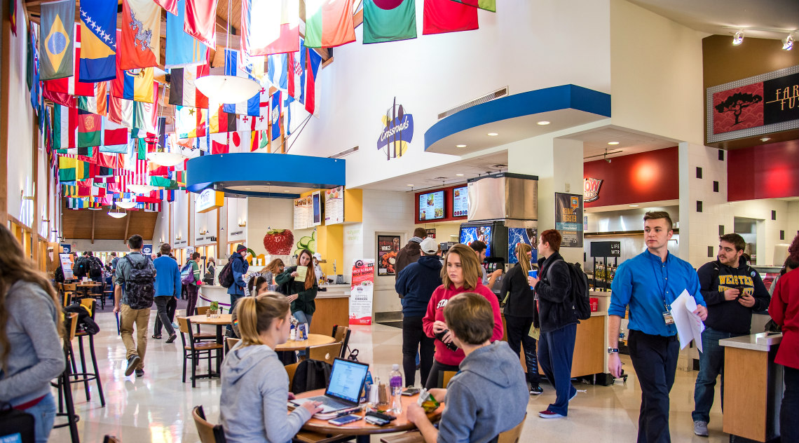 University Center hub with international flags overhead and students at tables and walking through hallway