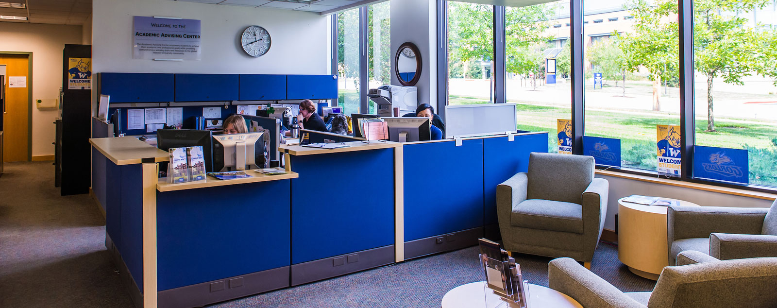 View of the Academic Advising center's reception area with blue front desk and several soft chairs in front of it.