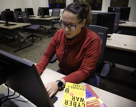 A female student working intently on a laptop with a Cyber War book by her side. 
