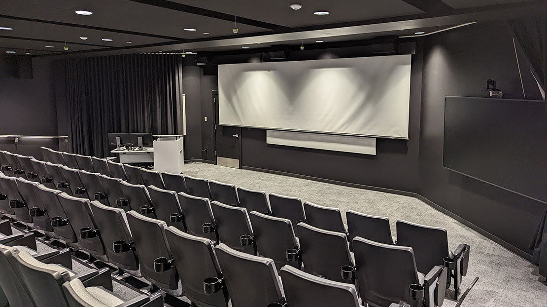 Gray screening room with three rows of theatre seats and large white widescreen, two large LED TV monitors on either side.