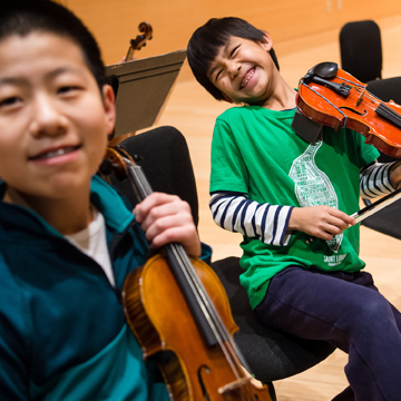 two young boys seated and holding violins and grinning