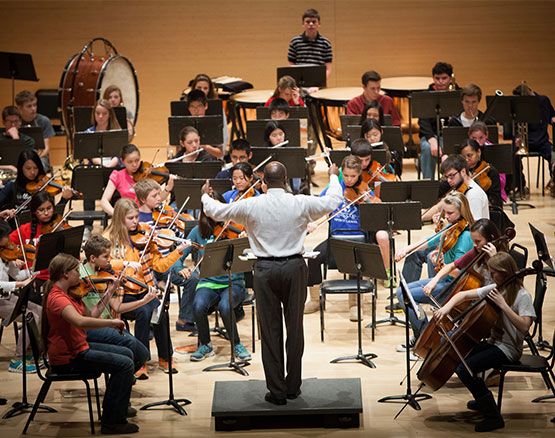 Conductor stands with arms out in front of orchestra of children with stringed instruments