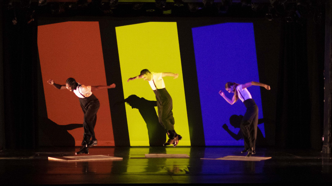 Three dancers set against a red, yellow, and blue backdrop, dancing in unison.