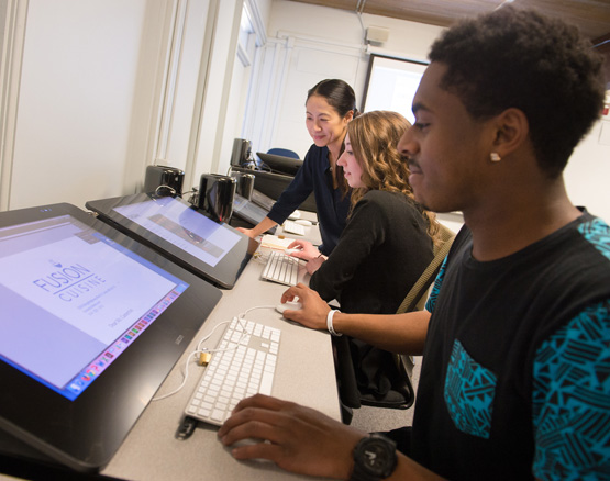 Students working on graphic designs on desk computers with professor