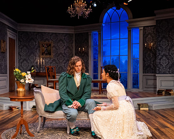Actor wearing green sits on period chaise, actress in cream dress kneels in front.