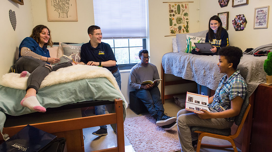 Group of six students in dorm room sitting in chair, on desk, on floor, and on beds all talking