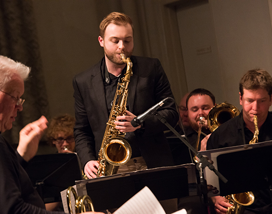 Two male students playing saxophones and one playing trombone with a director