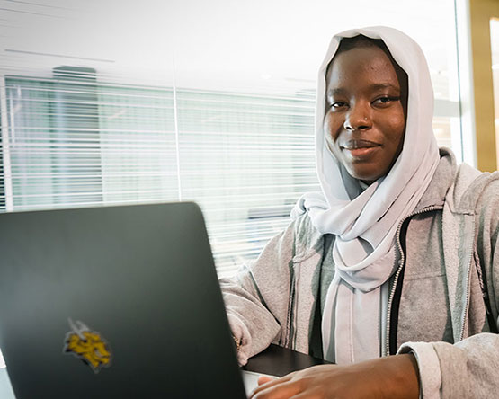 One female student smiling while sitting at a laptop with a Gorlok sticker on the cover. 
