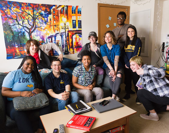 Diverse group of students sit on and around a couch in a dorm apartment, posing for camera.
